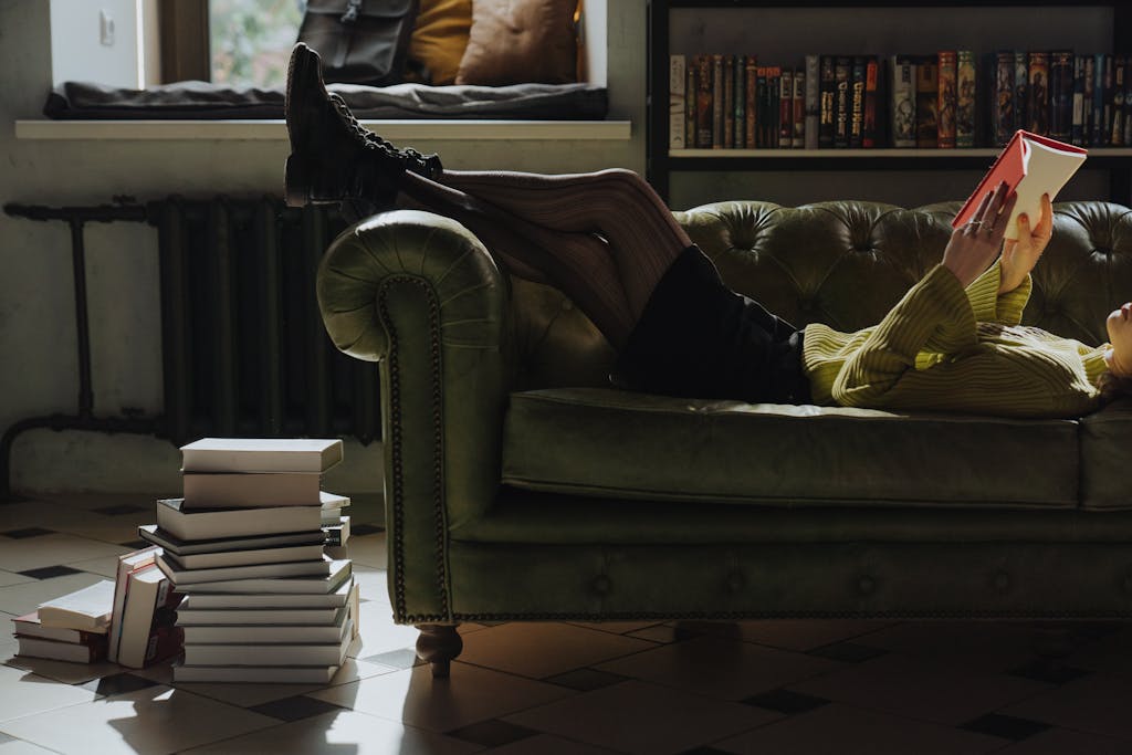 Person relaxing on a sofa reading a book indoors, surrounded by a stack of books.